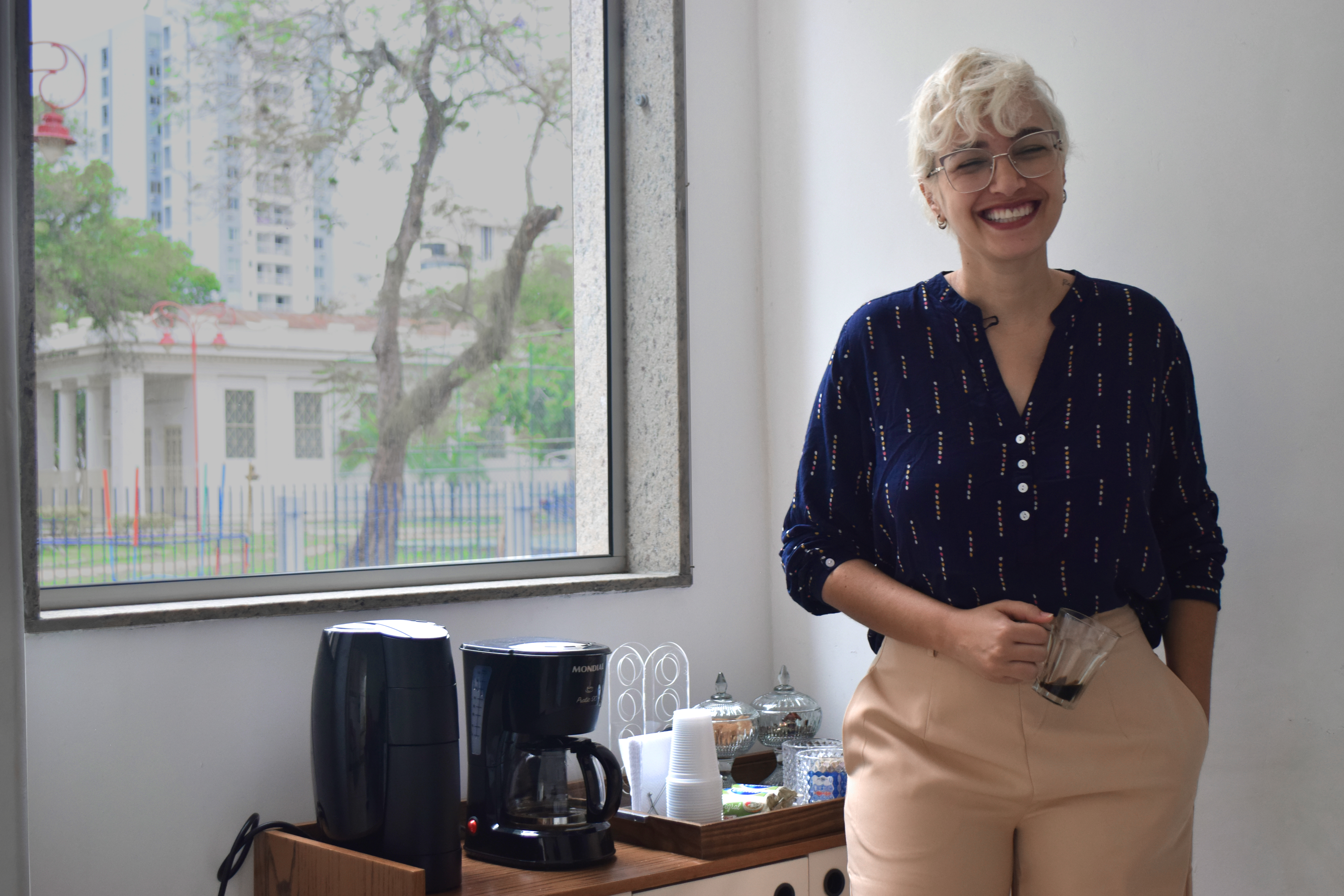 Psicóloga Maria Renata Reis sorrindo com uma xícara de café na mão ao lado de uma janela