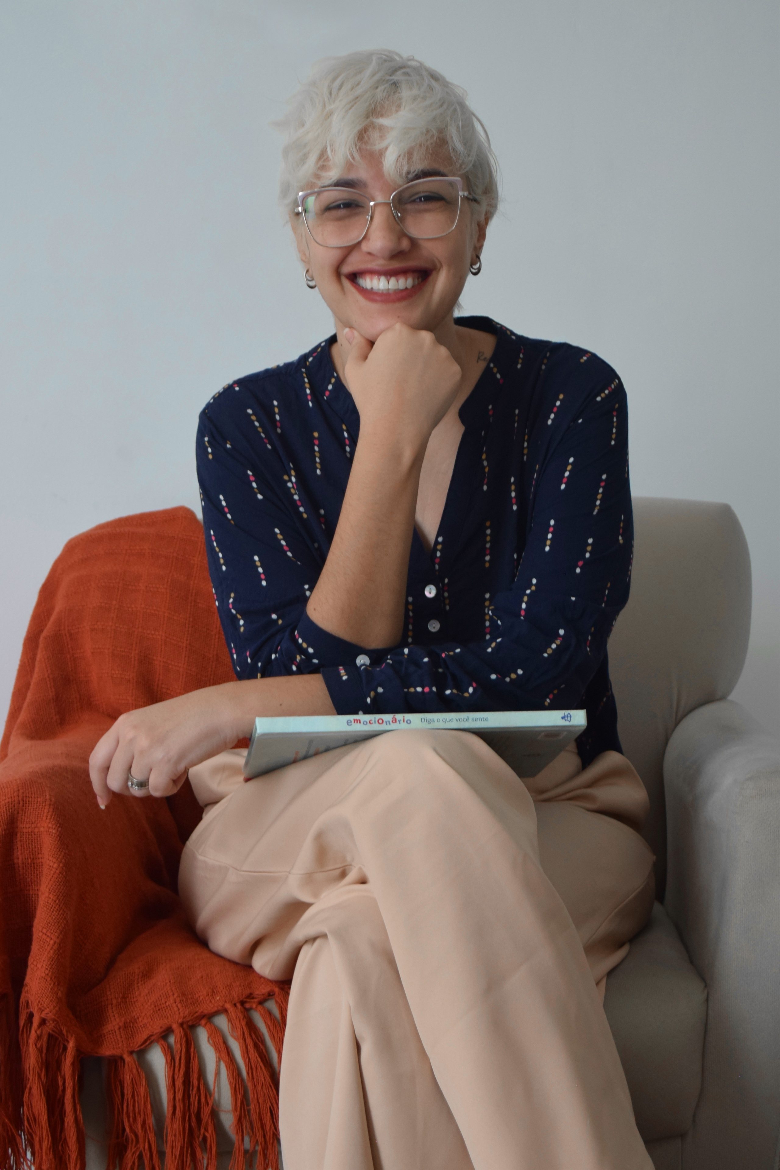 Psicóloga Maria Renata Reis sentada em uma poltrona sorrindo, com a mão direita no queixo, a esquerda sobre um livro repousado em suas pernas.